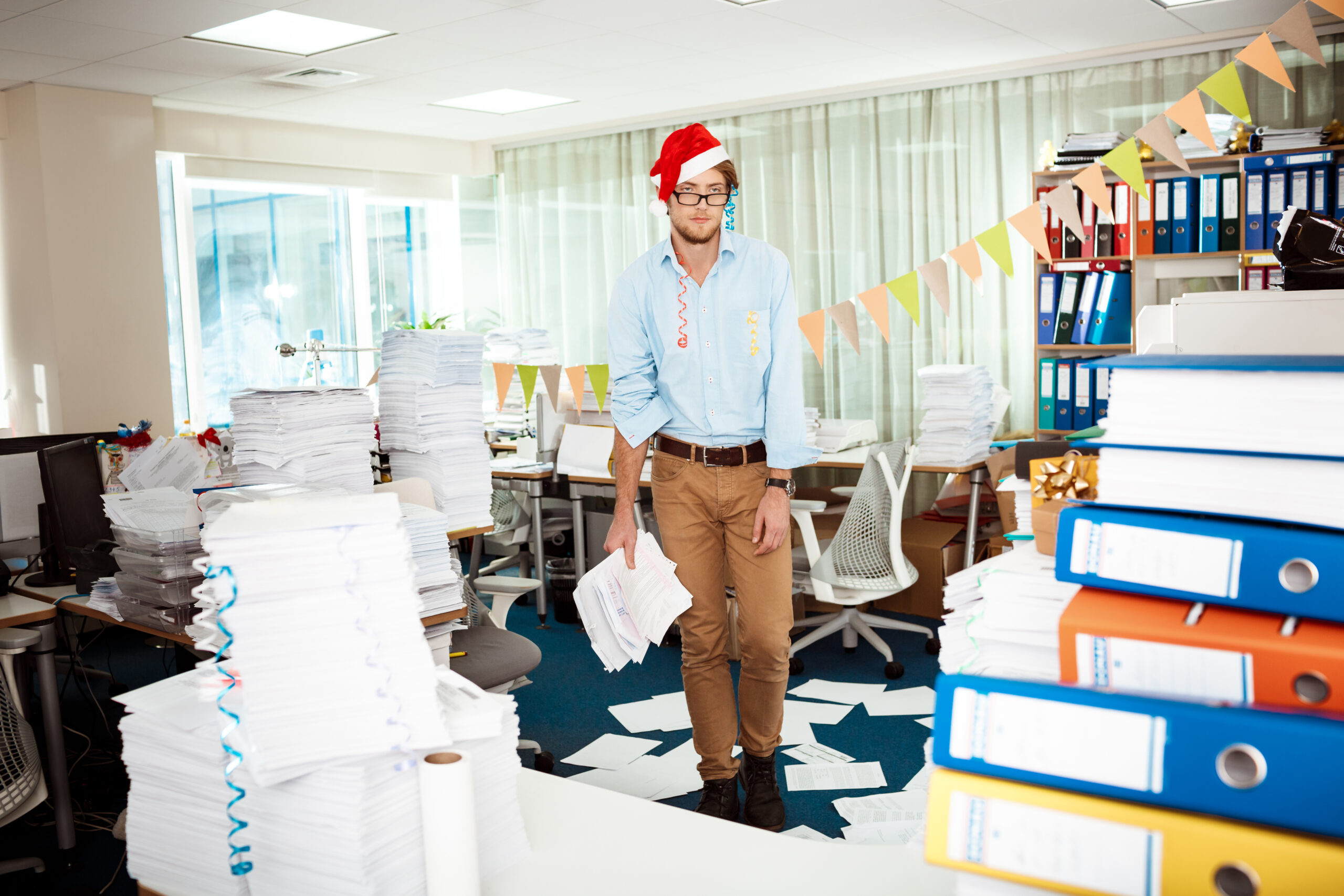 Tired young handsome businessman in red santa claus hat working in office among papers on christmas day.