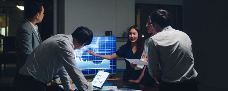 Four people in business attire discuss data shown on a digital screen and papers in a dimly lit office meeting room, carefully reviewing strategies for secure IT asset disposal to ensure sensitive information remains protected.