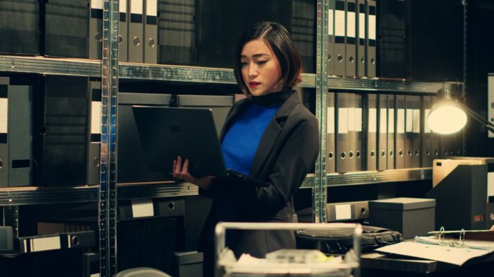 A woman in business attire holds a laptop, representing a potential candidate for laptop buyback, and stands in an office with shelves of files and boxes.