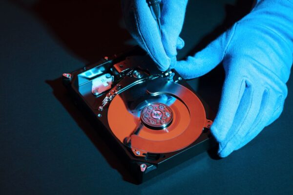 Gloved hands use a screwdriver to work on the exposed internal components of a hard disk drive, illuminated with blue and red lighting—demonstrating the meticulous process involved in professional data destruction and media destruction services.