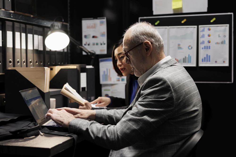 Two people work at a desk with documents and a laptop, surrounded by binders and charts pinned on the wall in an office setting, carefully discussing their company’s hard drive disposal service to ensure secure data destruction.