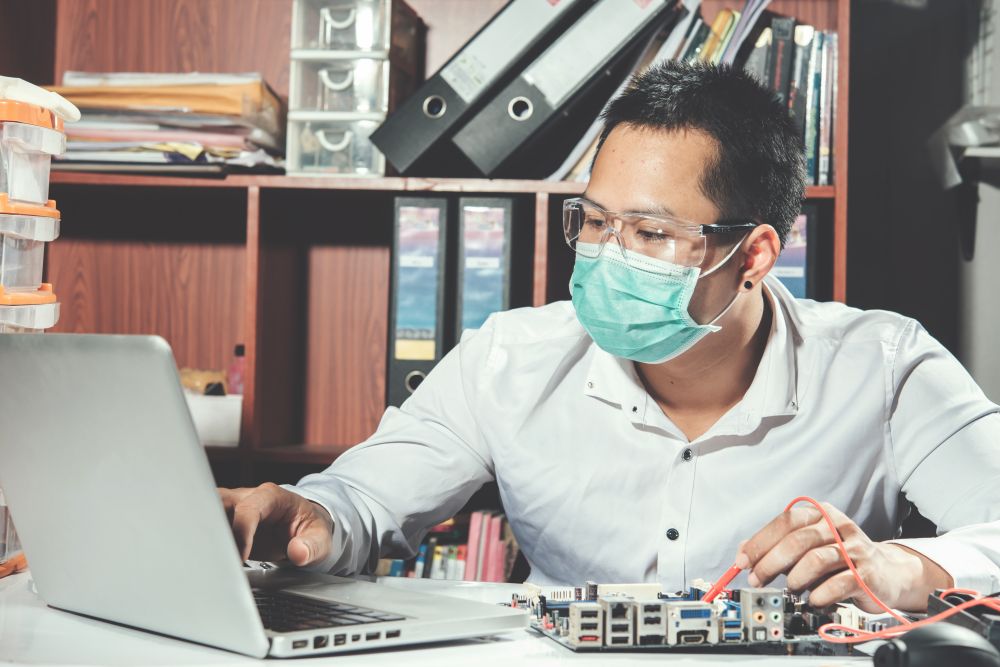 Man wearing a face mask and safety goggles works on a laptop and electronics circuit board at a desk with shelves and binders in the background, reflecting the careful attention to detail practiced by IT asset recycling companies.