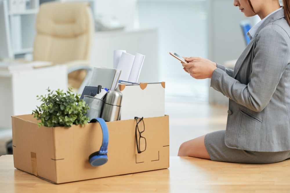 A person in business attire sits by a desk, using a phone. In front of them is a cardboard box filled with office items, suggesting a recent move or job change—a scene often seen when partnering with an ITAD company specializing in secure IT asset disposal.